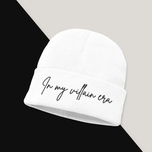 May include: White beanie with the text "In my villain era" in black cursive script.