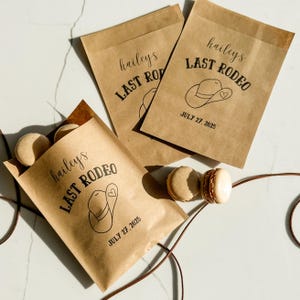 May include: Three brown paper bags with the text "hailey's LAST RODEO" and a cowboy hat graphic. The date "JULY 27, 2025" is printed below. Macarons are visible near the bags. A brown string is also visible.