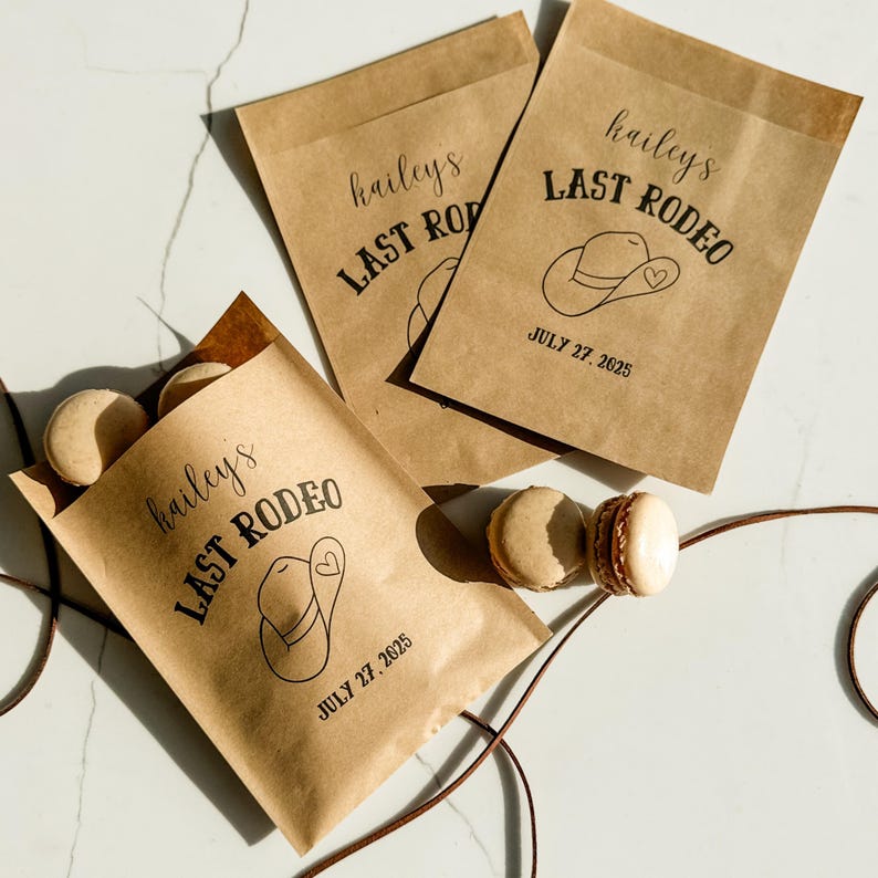 May include: Three brown paper bags with the text "hailey's LAST RODEO" and a cowboy hat graphic. The date "JULY 27, 2025" is printed below. Macarons are visible near the bags. A brown string is also visible.
