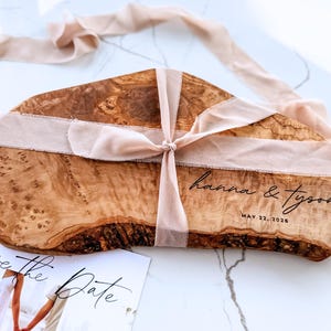 Puede incluir: Una tabla de servir de madera con acabado marrón claro, atada con una cinta color rubor. La tabla está grabada con los nombres "hanna & tyson" y la fecha "MAY 22, 2026". Se ve una tarjeta "Save the Date".