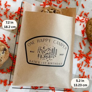 May include: A brown paper treat bag measuring 7.2 inches by 5.2 inches, featuring a black graphic of mountains, trees, and a tent with the text ONE HAPPY CAMPER, MAY 2027, and KATE'S 1ST BIRTHDAY. A chocolate chip cookie is visible inside the bag.