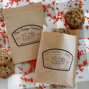 May include: Two rectangular brown paper bags featuring a black printed graphic of a mountain landscape, a tent, and trees. One bag reads One Happy Camper, July 2027, Harrison's First Birthday. The other reads One Happy Camper, May 2027, Kate's First Birthday.
