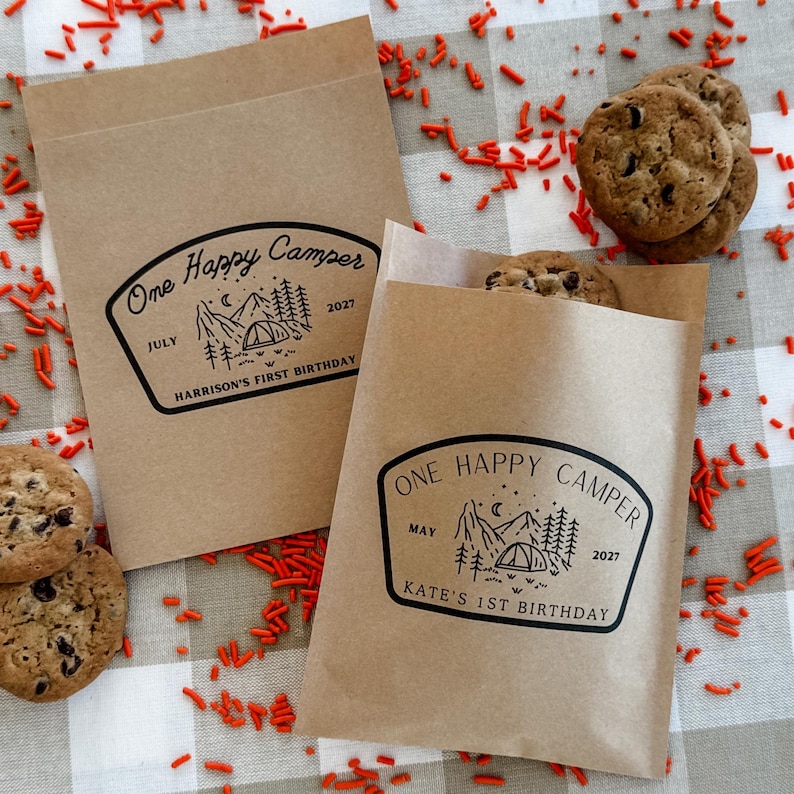 May include: Two rectangular brown paper bags featuring a black printed graphic of a mountain landscape, a tent, and trees. One bag reads One Happy Camper, July 2027, Harrison's First Birthday. The other reads One Happy Camper, May 2027, Kate's First Birthday.