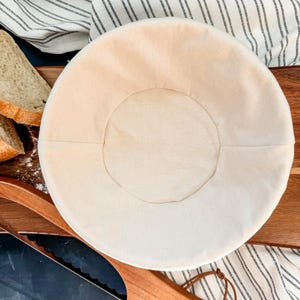 May include: A white linen proofing basket for baking bread. The basket is round and has a smooth, even surface. It is sitting on a wooden cutting board.