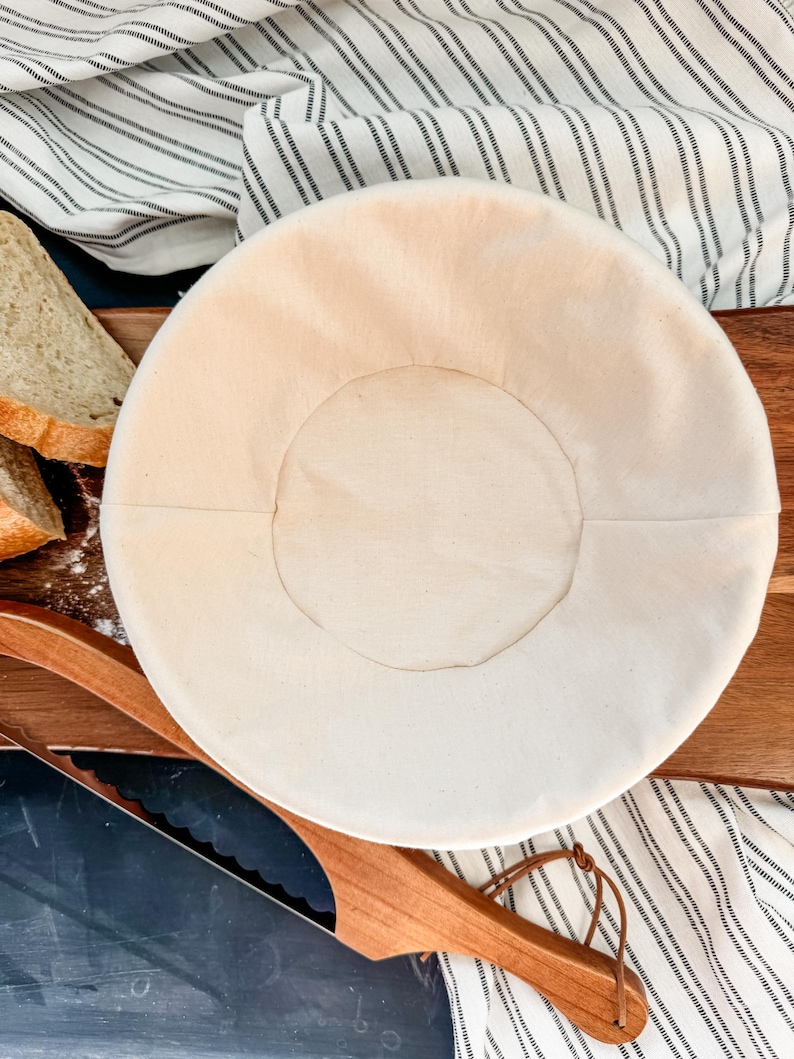 May include: A white linen proofing basket for baking bread. The basket is round and has a smooth, even surface. It is sitting on a wooden cutting board.