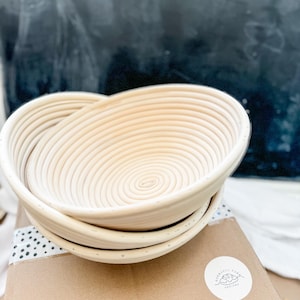 May include: A set of two round, woven, light brown bread proofing baskets. The baskets are stacked on top of each other. The baskets are on a brown cardboard box with a white sticker that says "Curated Pantry Designs".