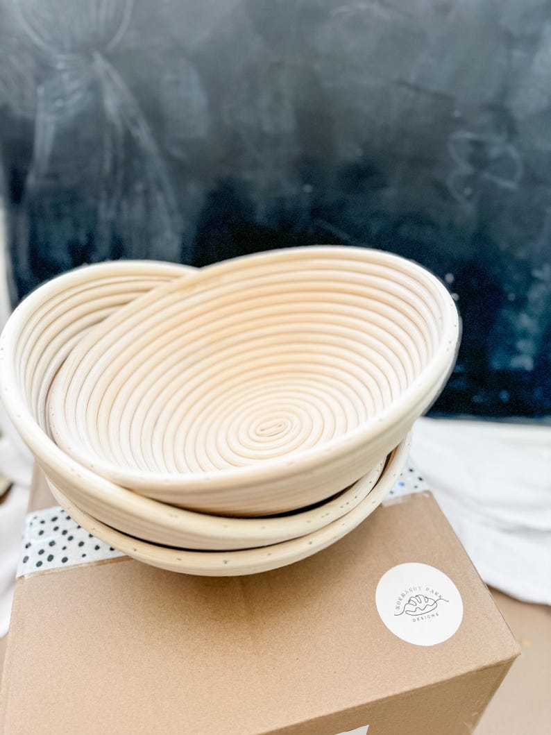May include: A set of two round, woven, light brown bread proofing baskets. The baskets are stacked on top of each other. The baskets are on a brown cardboard box with a white sticker that says "Curated Pantry Designs".