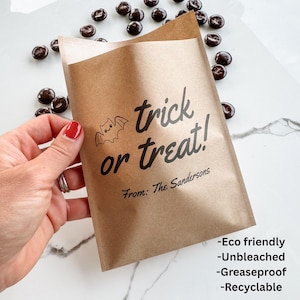 May include: A brown paper treat bag featuring a black printed bat icon and the text "trick or treat!" followed by "From: The Sandersons". The bag is surrounded by small dark chocolate candies, and the text "Eco friendly", "Unbleached", "Greaseproof", and "Recyclable" is printed in the bottom right corner.