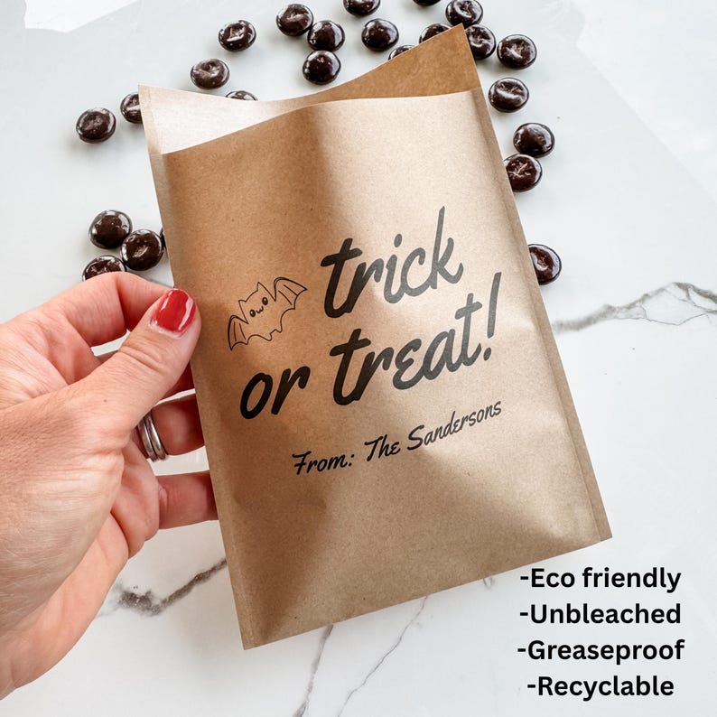 May include: A brown paper treat bag featuring a black printed bat icon and the text "trick or treat!" followed by "From: The Sandersons". The bag is surrounded by small dark chocolate candies, and the text "Eco friendly", "Unbleached", "Greaseproof", and "Recyclable" is printed in the bottom right corner.