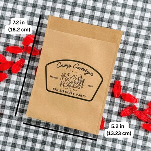 May include: A rectangular brown paper bag featuring a black printed graphic of a tent, mountains, trees, and stars. The text reads Camp Camryn, March 2026, 6th Birthday Party. The bag measures 7.2 inches in height and 5.2 inches in width.
