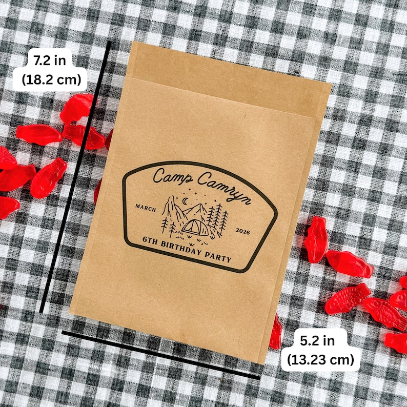 May include: A rectangular brown paper bag featuring a black printed graphic of a tent, mountains, trees, and stars. The text reads Camp Camryn, March 2026, 6th Birthday Party. The bag measures 7.2 inches in height and 5.2 inches in width.