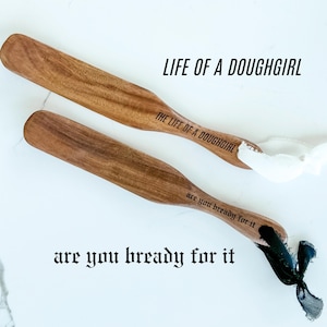 May include: Two wooden spatulas with the text "LIFE OF A DOUGHGIRL" and "THE LIFE OF A DOUGHGIRL" printed on them. The spatulas also have the text "are you bready for it" printed on them. One spatula has a white ribbon, and the other has a black ribbon.