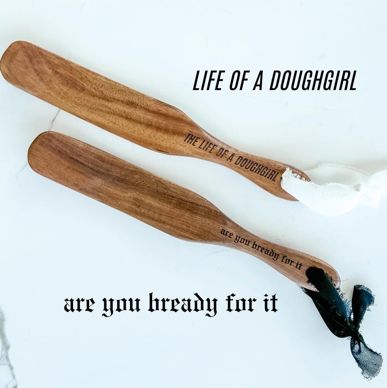 May include: Two wooden spatulas with the text "LIFE OF A DOUGHGIRL" and "THE LIFE OF A DOUGHGIRL" printed on them. The spatulas also have the text "are you bready for it" printed on them. One spatula has a white ribbon, and the other has a black ribbon.