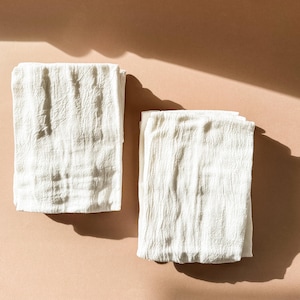 Puede incluir: Dos toallas de cocina de algod&oacute;n blanco dobladas cuidadosamente sobre una superficie marr&oacute;n claro.