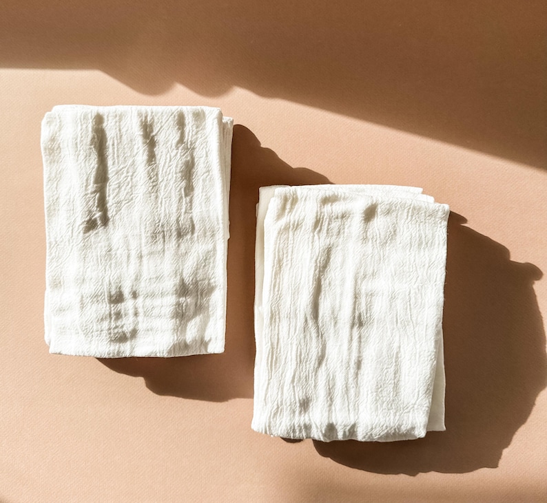 Puede incluir: Dos toallas de cocina de algod&oacute;n blanco dobladas cuidadosamente sobre una superficie marr&oacute;n claro.
