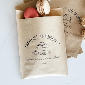 Op de afbeelding: Kraftpapieren cadeauzakjes met de tekst "FRESH OFF THE MARKET!" en een aardbeienillustratie. De zakjes bevatten twee macarons, één rood en één bruin. De tekst "Welcome to Kate's Bridal Shower!" staat ook op de zak.