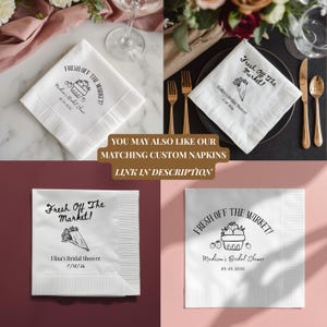 May include: White paper napkins with black text and illustrations. The napkins feature the phrase "Fresh Off The Market!" and bridal shower details. The image also includes gold forks, knives, and spoons, and floral arrangements.