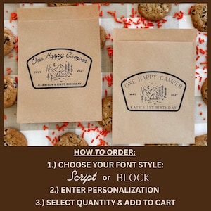 May include: Two rectangular brown paper bags featuring a black ink stamp design of a mountain landscape with a tent, trees, and the text One Happy Camper. One bag reads July 2027 Harrison's First Birthday, the other May 2027 Kate's 1st Birthday.