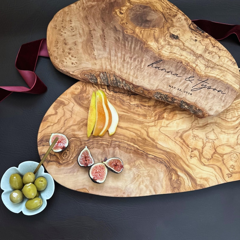 May include: Two olive wood serving boards with natural edges. One board has the inscription "hanna & tyson" and "May 22, 2026". The boards are decorated with sliced figs, pears, and olives in a blue floral dish. A burgundy ribbon is draped nearby.