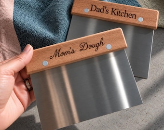 Peut inclure: Deux racloirs à pâte en acier inoxydable avec des manches en bois. Un manche est gravé avec le texte 'Dad's Kitchen' et l'autre avec 'Mom's Dough'. Chaque manche est fixé à la lame en métal par deux rivets métalliques argentés.