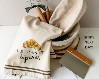 May include: A beige linen tea towel with a watercolor croissant and the words "LE PETIT déjeuner". Includes a bread lame, dough scraper, a wire whisk, and a banneton proofing basket. The set is designed for baking.