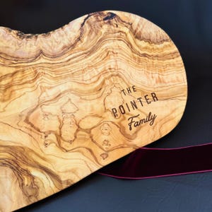 May include: A wooden serving board with a natural wood grain pattern. The board has the text "THE POINTER Family" engraved on it. A burgundy ribbon is draped across the bottom of the board.