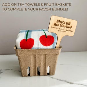 May include: A gift basket containing a cherry-themed tea towel and a wooden sign. The tea towel features two large red cherries with green stems on a blue and white striped background. The wooden sign reads "She's Off the Market!" and "OLIVIA'S BRIDAL SHOWER 7.12.2025".