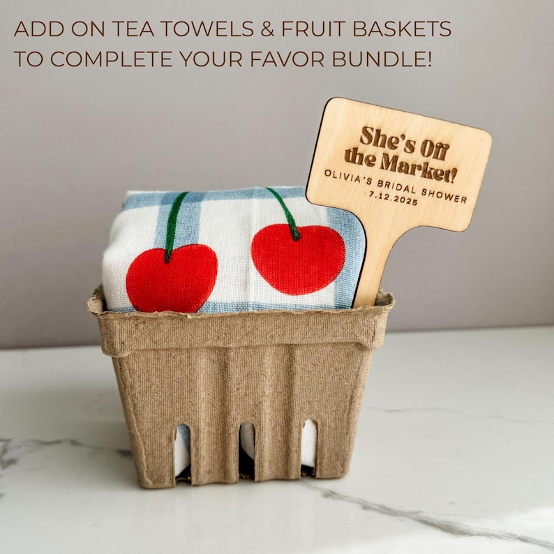 May include: A gift basket containing a cherry-themed tea towel and a wooden sign. The tea towel features two large red cherries with green stems on a blue and white striped background. The wooden sign reads "She's Off the Market!" and "OLIVIA'S BRIDAL SHOWER 7.12.2025".
