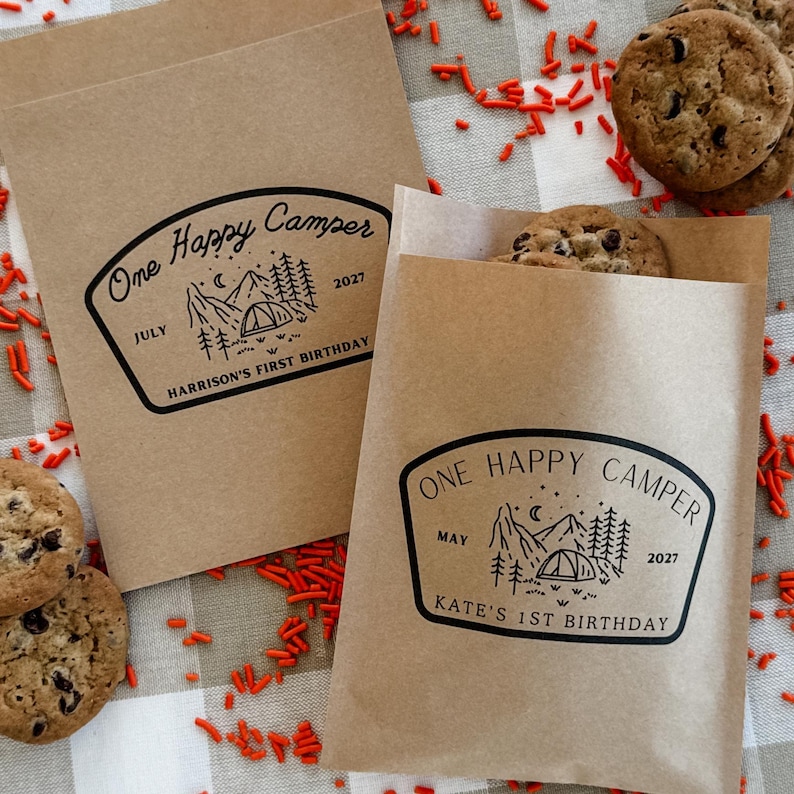 May include: Two brown paper bags featuring a black printed graphic of a tent, mountains, and trees with the text One Happy Camper, July 2027, Harrison's First Birthday and May 2027, Kate's 1st Birthday, surrounded by chocolate chip cookies.