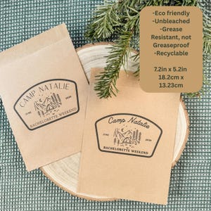 Pode incluir: Duas sacolas de papel retangulares marrom-claras com um gr&aacute;fico impresso em preto de uma paisagem de montanha, uma barraca e o texto Camp Natalie Bachelorette Weekend June 2026. As sacolas medem 18,2 cm por 13,23 cm e s&atilde;o recicl&aacute;veis.