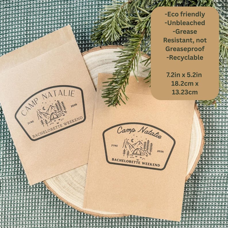 Pode incluir: Duas sacolas de papel retangulares marrom-claras com um gr&aacute;fico impresso em preto de uma paisagem de montanha, uma barraca e o texto Camp Natalie Bachelorette Weekend June 2026. As sacolas medem 18,2 cm por 13,23 cm e s&atilde;o recicl&aacute;veis.