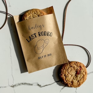May include: A brown paper bag with the text "hailey's LAST RODEO" and a cowboy hat illustration. The date "JULY 27, 2025" is printed below. Two cookies are visible, one inside the bag and one outside.