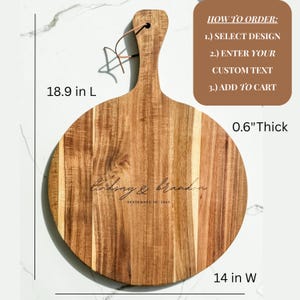 Puede incluir: Una tabla de cortar redonda de madera con asa, de 48 cm de largo y 36 cm de ancho, y 1,5 cm de grosor. La tabla presenta los nombres grabados "Lindsay & Brandon" y la fecha "SEPTEMBER 26, 2021". Una presilla de cuero est&aacute; unida al asa.
