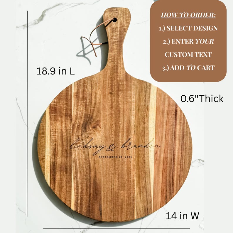 Puede incluir: Una tabla de cortar redonda de madera con asa, de 48 cm de largo y 36 cm de ancho, y 1,5 cm de grosor. La tabla presenta los nombres grabados "Lindsay & Brandon" y la fecha "SEPTEMBER 26, 2021". Una presilla de cuero est&aacute; unida al asa.