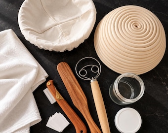 May include: A sourdough bread baking set including a round rattan proofing basket, a fabric liner, a wooden lame with replacement blades, a wooden dough whisk, a wooden spatula, a clear glass jar with a white lid, and a white cotton cloth.