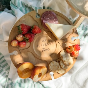 Portable charcuterie board and small wine table combo, featuring two wine glasses and a bottle of wine on a grassy picnic setting.