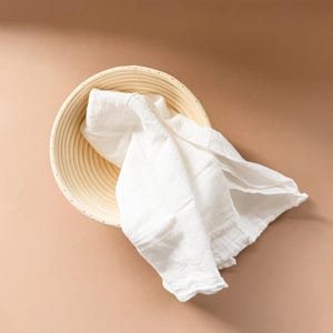 Puede incluir: Un pa&ntilde;o de lino blanco drapeado sobre una cesta redonda de mimbre para la fermentaci&oacute;n del pan. La cesta est&aacute; hecha de madera natural de color claro y tiene una superficie texturizada.