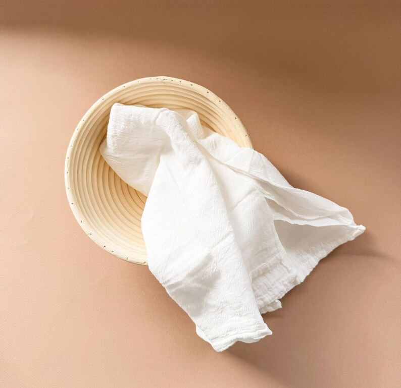 Puede incluir: Un pa&ntilde;o de lino blanco drapeado sobre una cesta redonda de mimbre para la fermentaci&oacute;n del pan. La cesta est&aacute; hecha de madera natural de color claro y tiene una superficie texturizada.