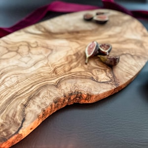 May include: A wooden serving board with a natural edge, featuring a light brown and tan wood grain pattern. The board is adorned with sliced figs and a burgundy ribbon. This rustic board is ideal for serving appetizers or charcuterie.