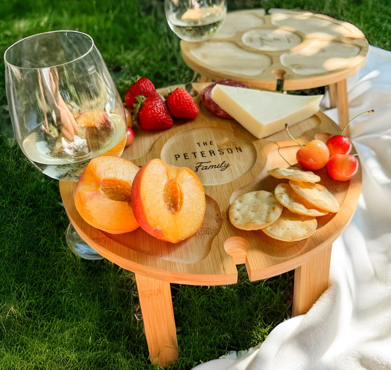 Foldable personalized wine table with built-in wine glass holders, set outdoors for a picnic with wine and cheese.