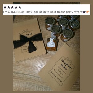 May include: A stack of light brown paper cards with black text reading Bonjour B&eacute;b&eacute;, Welcome to Ashley's Baby Shower, DECEMBER 27, 2023, tied with a black frayed ribbon, alongside several small glass jars filled with amber-colored spread.
