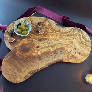 May include: Two olive wood serving boards with personalized text. One board is engraved with "Hanna & Tyson" and the date "May 23, 2020." The other board is engraved with "The Pointer Family." A small bowl of olives and figs are on the boards.