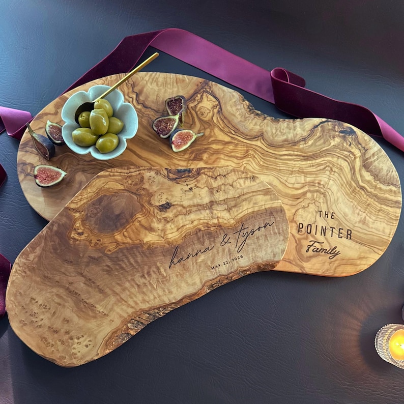 May include: Two olive wood serving boards with personalized text. One board is engraved with "Hanna & Tyson" and the date "May 23, 2020." The other board is engraved with "The Pointer Family." A small bowl of olives and figs are on the boards.