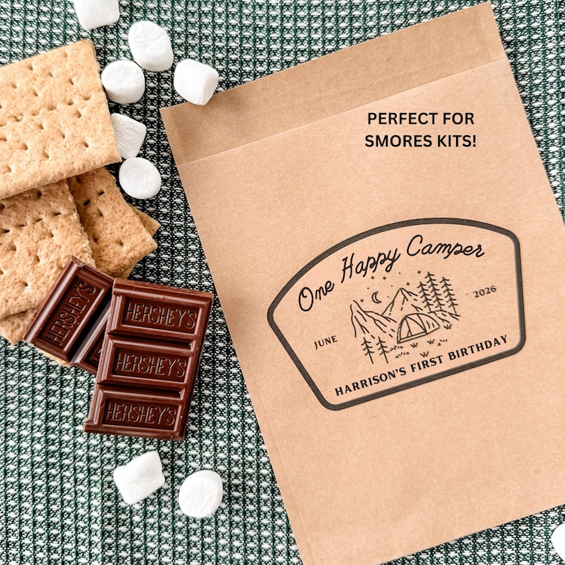 May include: A brown paper bag with a black graphic print featuring a tent, mountains, trees, and the text "One Happy Camper", "JUNE", "2026", and "HARRISON'S FIRST BIRTHDAY". The bag also includes the text "PERFECT FOR SMORES KITS!" next to graham crackers and Hershey's chocolate bars.