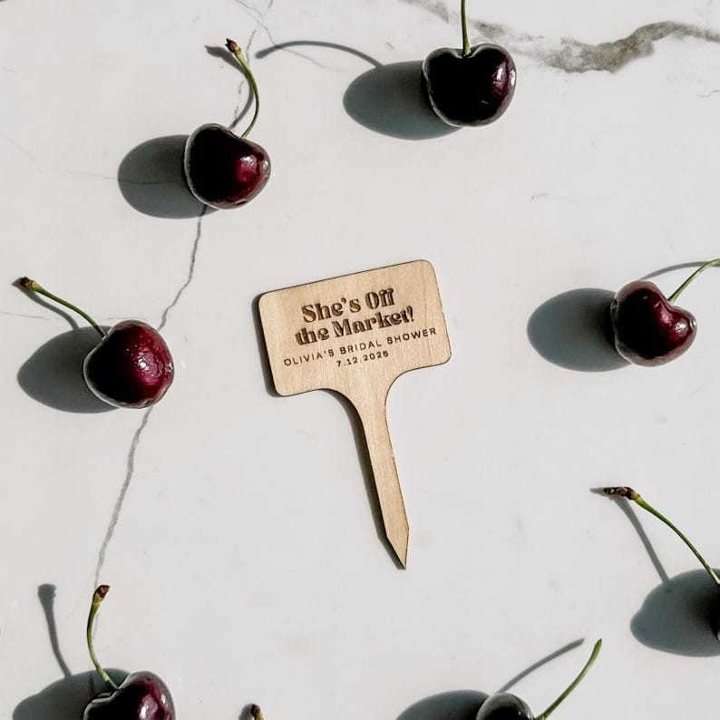 May include: A wooden sign with the text "She's Off the Market!" and "Olivia's Bridal Shower 7.12.2028" is surrounded by fresh cherries. The cherries are a deep red color with green stems, arranged on a white surface.