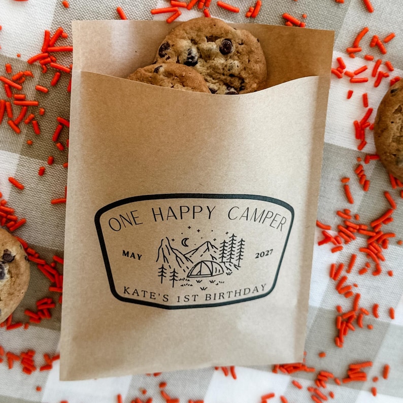 May include: A brown paper treat bag containing chocolate chip cookies. The bag features a black printed graphic of a mountain, tent, and trees with the text ONE HAPPY CAMPER, MAY 2027, and KATE'S 1ST BIRTHDAY. The bag is surrounded by orange sprinkles.