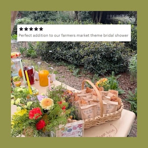 May include: A woven wicker basket filled with small brown paper bags containing pastries, each labeled with the text Fresh Off The Market, positioned next to a colorful floral arrangement, glass pitchers of juice, and tall taper candles.