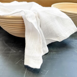 Puede incluir: Un pa&ntilde;o blanco drapeado sobre una cesta redonda tejida con un acabado de madera natural. La cesta est&aacute; sobre una superficie negra.