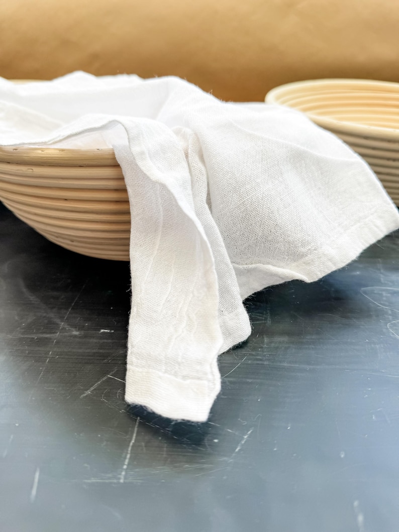 Puede incluir: Un pa&ntilde;o blanco drapeado sobre una cesta redonda tejida con un acabado de madera natural. La cesta est&aacute; sobre una superficie negra.