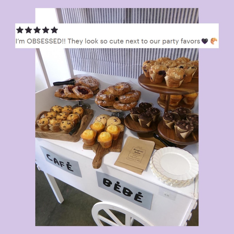 Pode incluir: Um carrinho branco com a etiqueta CAF&Eacute; B&Eacute;B&Eacute; exibe uma variedade de produtos de padaria, incluindo muffins, past&eacute;is e biscoitos em bandejas de madeira e suportes de v&aacute;rios n&iacute;veis. Inclui pratos brancos e sacos de papel com o texto Bonjour b&eacute;b&eacute;.
